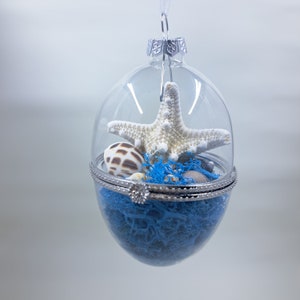 May include: A clear glass ornament with a silver hinged lid, filled with blue moss, seashells, and a white starfish.