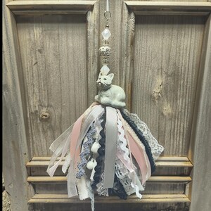 May include: A hanging ornament featuring a light gray ceramic cat figurine perched atop a cluster of layered ribbons and lace. The ribbons are in shades of pale pink, black, white, and gray, accented by a vertical string of clear and silver beads.