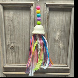 May include: A decorative hanging ornament featuring a vertical stack of five round silicone beads in pink, yellow, turquoise, purple, and lime green, topped by a cream-colored bell shape with a cluster of multi-colored fabric ribbons and trim.