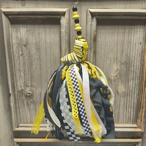 May include: A hanging ornament featuring a yellow and black striped caterpillar figure attached to a cluster of ribbons. The ribbons are yellow, black, and white, with various textures including checkered patterns, glitter, and solid colors.