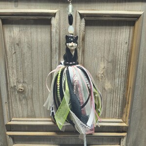 May include: A decorative hanging ornament featuring a black and white ceramic cat figurine perched atop a multi-textured tassel. The tassel consists of various ribbons in black, white, lime green, and pink, accented with lace, sequins, and beads.
