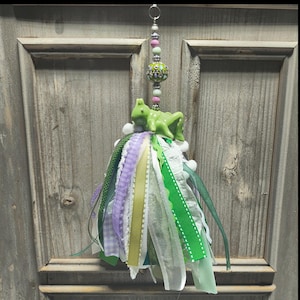 May include: A hanging ornament featuring a green grasshopper figurine atop a cluster of layered ribbons in shades of green, white, and lavender. The ornament is suspended by a vertical strand of decorative beads in pink, white, and patterned green.