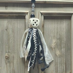 May include: A decorative hanging ornament featuring a white six-sided die with black pips, suspended from a string of black and white beads. Below the die is a cluster of assorted ribbons, including black and white polka dots, lace, and fringe.