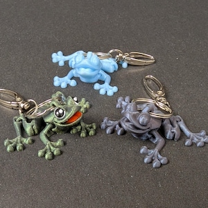 May include: Three frog keychains, one blue, one green, and one gray. Each frog is 3D printed and has a unique expression.