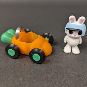 May include: A white rabbit wearing a blue helmet with black eyes and a pink nose is standing next to a small orange toy car shaped like a carrot. The car has green wheels and a silver handle.