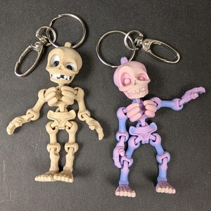 May include: Two skeleton keyrings. One is tan, the other is purple and blue. Both have articulated limbs and a metal keyring and clasp. The skeletons have detailed rib cages, skulls, and feet. The keyrings are on a dark grey surface.