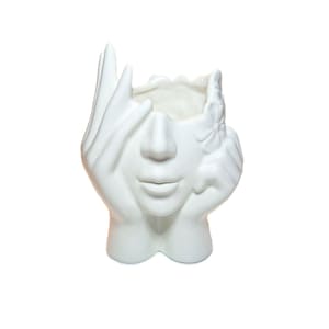 Op de afbeelding: Een witte keramische vaas in de vorm van een menselijk gezicht met handen eromheen. Eén hand bedekt het linkeroog, terwijl de andere hand tegen de rechterwang rust. Een kleine bloem is aan de rechterkant bij de slaapstreek gebeeldhouwd.