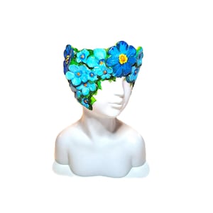 May include: A white ceramic head-shaped planter featuring a crown of sculpted blue flowers with yellow centers and green leaves. The floral arrangement wraps around the upper portion of the head, creating a vibrant contrast against the smooth white surface.