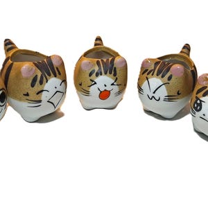 May include: Five ceramic cat-shaped planters with different facial expressions. The planters are tan with black stripes, pink ears, and white faces. Each planter has a unique, hand-painted expression.