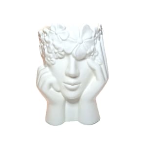 Könnte beinhalten: Ein mattweißer Keramik-Pflanztopf in Form eines menschlichen Kopfes, dessen Hände an den Wangen ruhen. Die Figur zeigt eine Krone aus modellierten Blumen über Stirn und Augen, mit einer glatten Gesichtsstruktur und einer hohlen Öffnung oben.