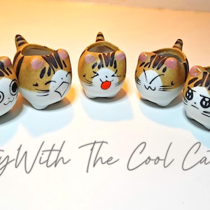 May include: Five ceramic cat-shaped planters with various facial expressions. The planters are tan with black stripes and white faces. The text "Party With The Cool Cats" is written below the planters.