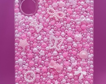May include: Pink phone case decorated with pink and white beads, pearls, and various embellishments. The case features a camera cutout and is adorned with bows, hearts, and star shapes. A decorative and stylish accessory.