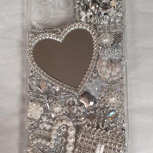 May include: A clear phone case adorned with silver rhinestones, pearls, and a heart-shaped mirror. The case is decorated with a small purse design and the letters "LV".