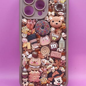 Puede incluir: Una funda de smartphone marrón decorada con un denso collage de dijes en miniatura que incluyen un oso de peluche, una dona, una taza de café, un cartón de leche, un ratón de dibujos animados y varias cuentas. Incluye texto como Love, M y MILK.