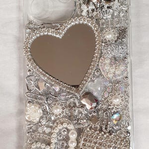 May include: Clear phone case embellished with silver rhinestones, pearls, and a heart-shaped mirror. Features a small purse design and various decorative elements, creating a glamorous aesthetic.