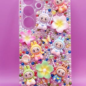 May include: A clear phone case decorated with a dense arrangement of multicoloured beads, pearls, and various charms including six Labubu figures, three flowers, two lollipops, two butterflies, and two chocolate donut charms with the text LABUBU at the bottom.