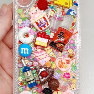 Könnte beinhalten: Transparente Handyhülle mit Miniatur-Nahrungsmittel- und Süßigkeiten-Anhängern verziert. Enthält eine Popcorn-Box, eine Wasserflasche, ein Stück Käse, einen Donut, eine Weinflasche, eine Kaffeetasse und andere farbenfrohe Verzierungen zum Thema Essen.