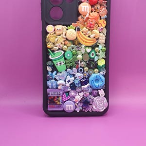 Puede incluir: Una funda de smartphone negra decorada con una densa disposición de abalorios en miniatura de colores en un patrón degradado. Los abalorios incluyen alimentos como un plátano, un té matcha, una bolsa de Haribo, M&Ms y varias flores.