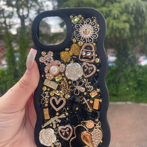 May include: Black phone case with a variety of decorative embellishments. The case features gold, silver, and clear rhinestones, a teddy bear, flowers, hearts, and other charms. The case has a glossy finish.
