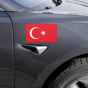 Könnte beinhalten: Ein rechteckiger roter Autoaufkleber mit der türkischen Flagge, mit einem weißen Halbmond und Stern. Der Aufkleber ist an der Seite eines dunkelgrauen Autos angebracht, in der Nähe des Vorderrads.