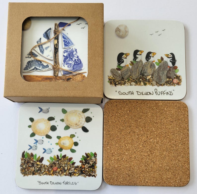 May include: A set of four square coasters. One coaster features a sailboat design with blue and white patterns. Another depicts puffins with the text "South Devon Puffins." The third shows turtles with the text "South Devon Turtles." The fourth is cork.