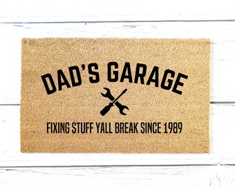 May include: A brown doormat with the text "Dad's Garage" in black lettering. Below the text is a wrench and screwdriver crossed and the text "Fixing stuff yall break since 1989".