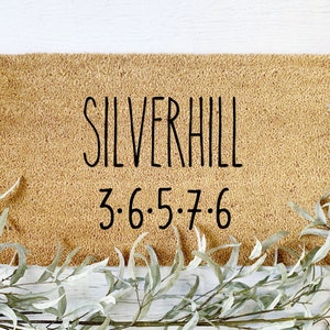 May include: A rectangular doormat with the word "SILVERHILL" in black script, and the numbers "3·6·5·7·6" below. The mat is a natural tan color, with a textured surface. Greenery surrounds the mat.