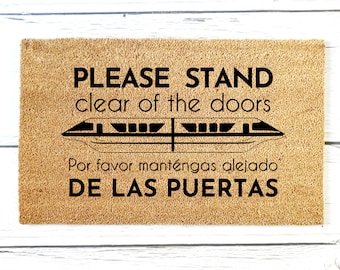 May include: A brown coir doormat with black text that reads "PLEASE STAND clear of the doors" and a black silhouette of a monorail. The text below the monorail reads "Por favor manténgas alejado DE LAS PUERTAS".