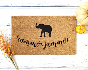 May include: A brown coir doormat with the text "rammer jammer" in black cursive lettering. A black silhouette of an elephant is centered above the text.