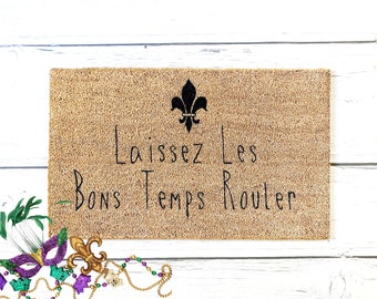 Peut inclure: Un paillasson en fibre de coco brun avec le texte "Laissez Les Bons Temps Rouler" en écriture noire. Un motif de fleur de lys noire est au-dessus du texte.