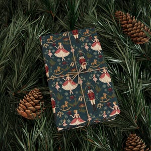 May include: A wrapped gift with a dark teal background and a repeating pattern of ballerinas, nutcrackers, rocking horses, and crowns. The gift is tied with twine and set against a backdrop of pine needles and pine cones.
