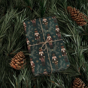 May include: A wrapped gift with a dark green background featuring a repeating pattern of nutcracker soldiers. The gift is tied with twine and rests on pine needles and pine cones, creating a festive holiday theme.