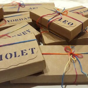 Puede incluir: Una pila de cajas de regalo marrones atadas con cuerda de colores. Cada caja tiene un nombre escrito en letras moradas. Los nombres incluyen Emma, Jordan, Leo, Liam, Violet, Tam, Yosef y Janry.