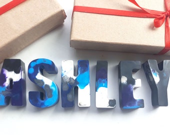 Puede incluir: El nombre "ASHLEY" escrito en letras de crayón azul, negro y blanco. Las letras están hechas de crayón derretido y tienen un aspecto marmoleado.