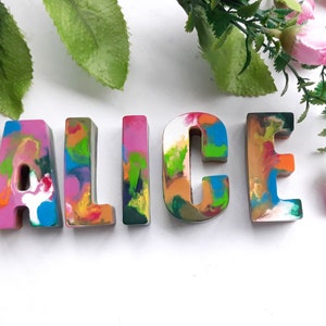 Puede incluir: Letras coloridas, multicolores y texturizadas que deletrean el nombre "ALICE" en una fuente caprichosa. Las letras est&aacute;n hechas de cera y tienen un aspecto &uacute;nico y hecho a mano.