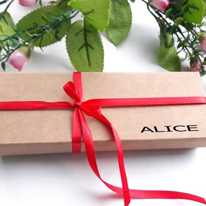 Puede incluir: Una caja de regalo marr&oacute;n con una cinta y un lazo rojo. La caja est&aacute; atada con una cinta roja y tiene el nombre "ALICE" impreso en la parte delantera.