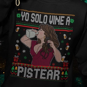 May include: Black sweatshirt with a festive design featuring the text "YO SOLO VINE A PISTEAR" and a woman drinking from a mug. The design is in green, red, and white. The person is holding a yellow camera and wearing a red headband.