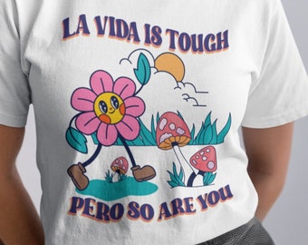 May include: White t-shirt with a retro graphic of a smiling flower with legs, mushrooms, and the text "LA VIDA IS TOUGH PERO SO ARE YOU" in purple and orange. The shirt is worn by a person.