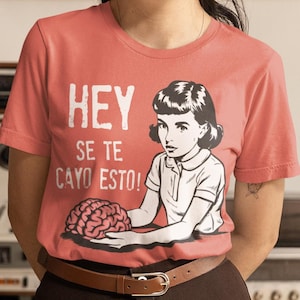 May include: A woman wearing a pink t-shirt with a graphic of a woman holding a brain and the text "HEY SE TE CAYO ESTO!"