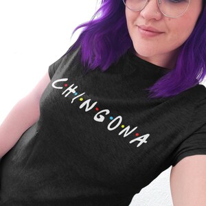 May include: A woman with purple hair and round glasses is wearing a black t-shirt with the word "CHINGONA" written in white letters with colorful dots.