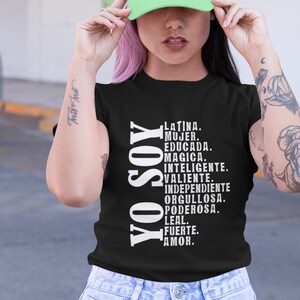 Puede incluir: Una camiseta negra con el texto "YO SOY" en blanco, seguido de una lista de palabras en espa&ntilde;ol que describen las cualidades de una mujer. La modelo lleva una gorra de b&eacute;isbol verde claro con el mismo texto en blanco.