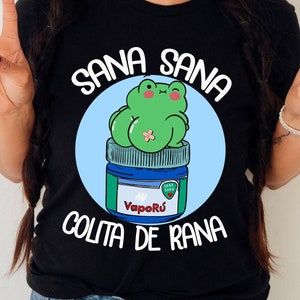 May include: Black t-shirt with a graphic featuring a green frog sitting on a blue and white jar of VapoRub. The text "Sana Sana" is arched above the frog, and "Colita de Rana" is below. The frog has a bandage and is winking.