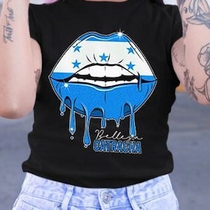 May include: A black t-shirt with a dripping blue and white lips design. The lips are decorated with white stars and the text "Pelleja Catracha" is printed below the lips.