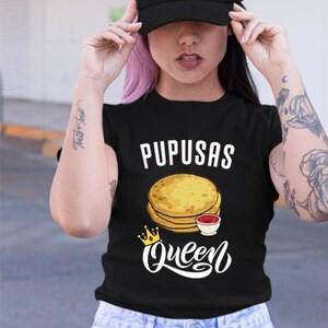 Puede incluir: Una camiseta negra con el texto "Pupusas Queen" y un gr&aacute;fico de una pila de pupusas con un lado de salsa. La camiseta la lleva puesta una persona con el pelo rosa y una gorra de b&eacute;isbol negra.