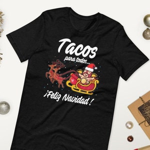 May include: Black t-shirt with a festive graphic featuring a pig in a Santa hat riding a sleigh pulled by reindeer. The text reads "Tacos para todos... &iexcl;Feliz Navidad!" in white, perfect for the holiday season.