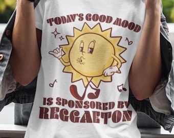 Könnte beinhalten: Weißes T-Shirt mit dem Text "TODAY'S GOOD MOOD IS SPONSORED BY REGGAETON" in Braun. Das Shirt zeigt eine Cartoon-Sonne mit Sonnenbrille und Noten. Die Person trägt eine Brille und eine Jeansjacke.