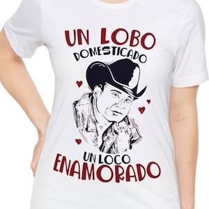 May include: White t-shirt featuring a graphic of a man in a cowboy hat with the text "UN LOBO DOMESTICADO UN LOCO ENAMORADO" in burgundy. The design includes small heart shapes.