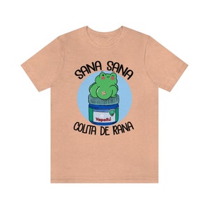 May include: A peach-colored t-shirt featuring a cartoon frog sitting on a blue and green jar of VapoR&uacute;. The text "SANA SANA COLITA DE RANA" is printed above and below the frog illustration.