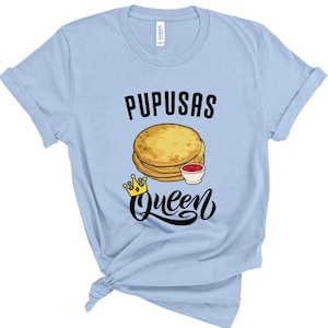 Puede incluir: Camiseta azul claro con el texto "Pupusas Queen" en negro. Una imagen de dibujos animados de pupusas con un lado de salsa est&aacute; por encima del texto. Una corona dorada est&aacute; debajo del texto.