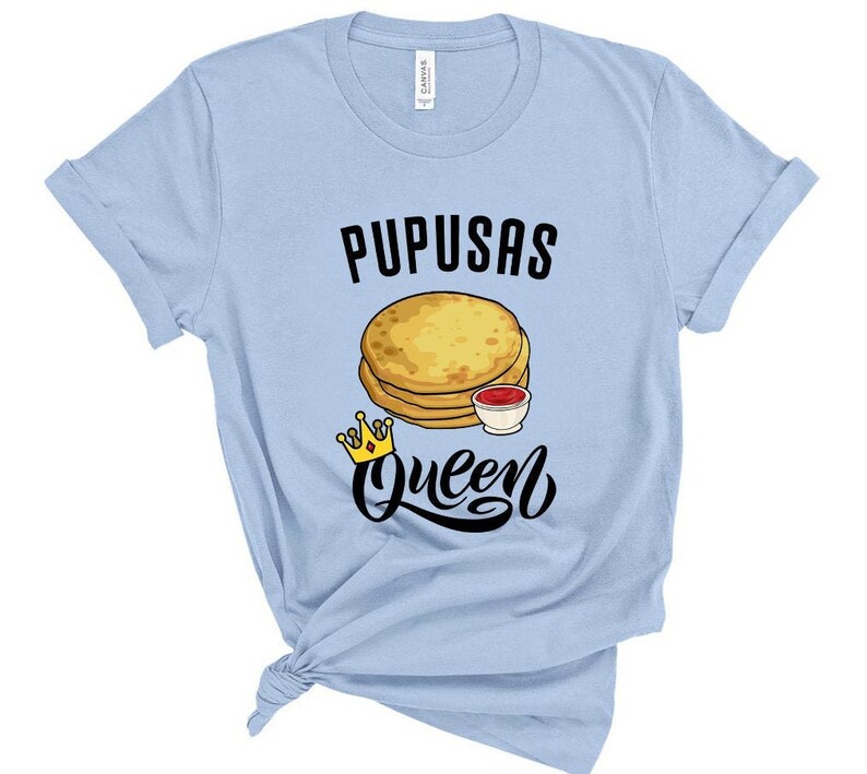 Puede incluir: Camiseta azul claro con el texto "Pupusas Queen" en negro. Una imagen de dibujos animados de pupusas con un lado de salsa est&aacute; por encima del texto. Una corona dorada est&aacute; debajo del texto.